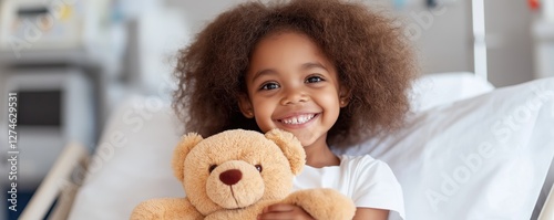 A young African American girl around four years old smiles shyly while clutching a teddy bear on her hospital bed. The spacious room provides a soothing atmosphere perfect for rest and recovery photo