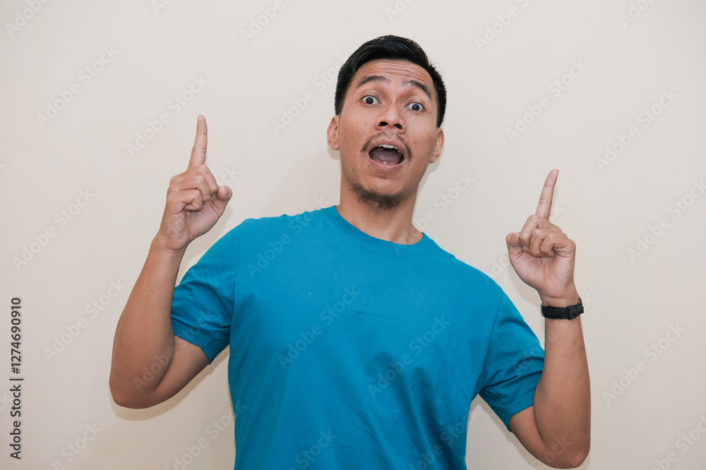 Fototapeta premium A Southeast Asian man in a turquoise green tshirt smiling and pointing up with both index fingers, in a white background. The image captures an exciting and curious expression 