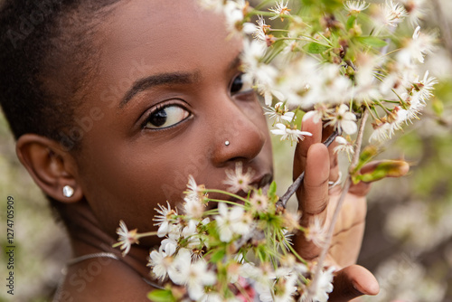 Beautiful young model with short hair and nose piercing gently touching and smelling delicate white cherry blossoms on a flowering tree branch in an orchard in springtime. Meditation, mental treatment
