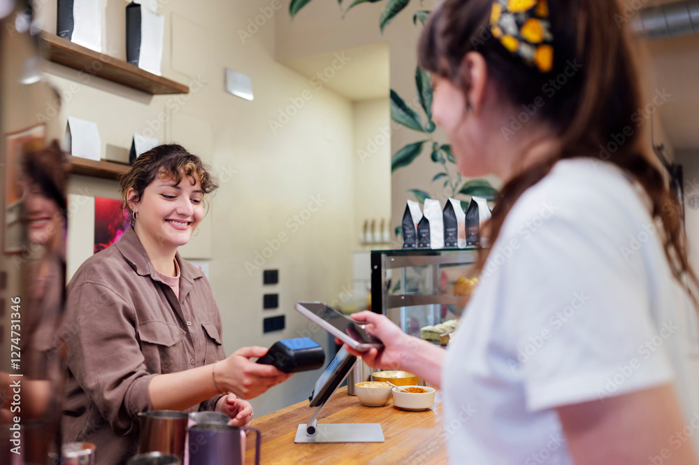 Fototapeta premium Smiling barista holding POS terminal while customer paying with smartphone using NFC technology
