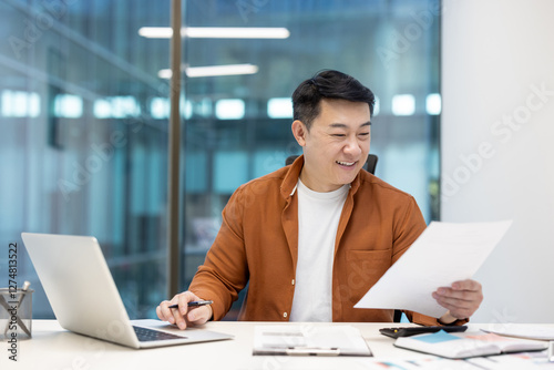 Successful joyful businessman investor doing paperwork, asian man working with documents reports and contracts. Man in casual clothes using laptop at work inside office.