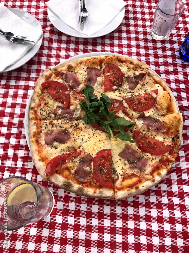 Pizza on a checkered tablecloth with tomato and ham. Freshly baked pizza with mozzarella tomato slices ham and herbs on a white plate. Pizza served on red and white checkered tablecloth Pizza on a checkered tablecloth with tomato and ham. Freshly baked pizza with mozzarella tomato slices ham and herbs on a white plate. Pizza served on red and white checkered tablecloth