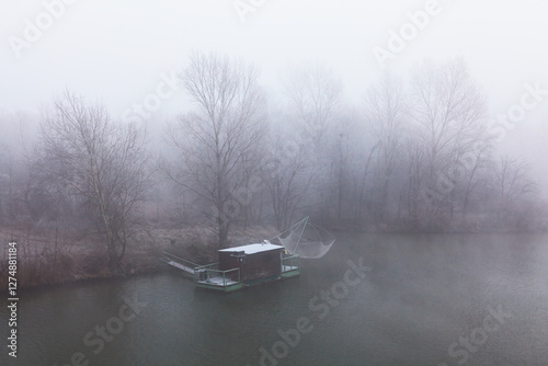 Obraz na płótnie Foggy river scene with a small fishing hut on a floating platform. Bare trees fade into the mist, creating a mysterious, tranquil atmosphere. Muted tones and soft light enhance the dreamlike mood w salonie