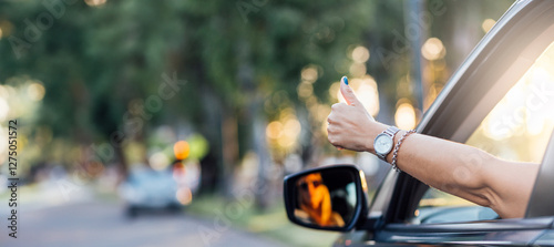A female driver puts her hand out the car window while driving and shows a thumbs up making an okay sign with her hand. Photo in banner format with copy space.
