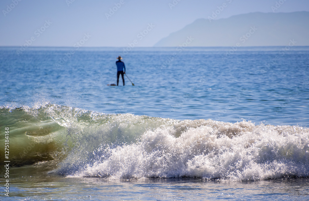 Man paddling out to sea on a clear day with crystal clear blue water and sky with a few clouds, sunny with hat and backpack - waves in the foreground Man paddling out to sea on a clear day with crystal clear blue water and sky with a few clouds, sunny with hat and backpack - waves in the foreground