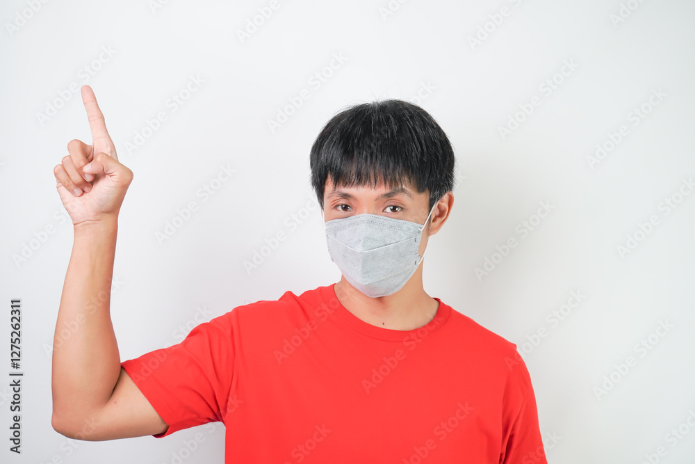 Young asian man in red wearing medical mask during virus outbreaks pandemic is pointing copy space above him, isolated over white background. Young asian man in red wearing medical mask during virus outbreaks pandemic is pointing copy space above him, isolated over white background.