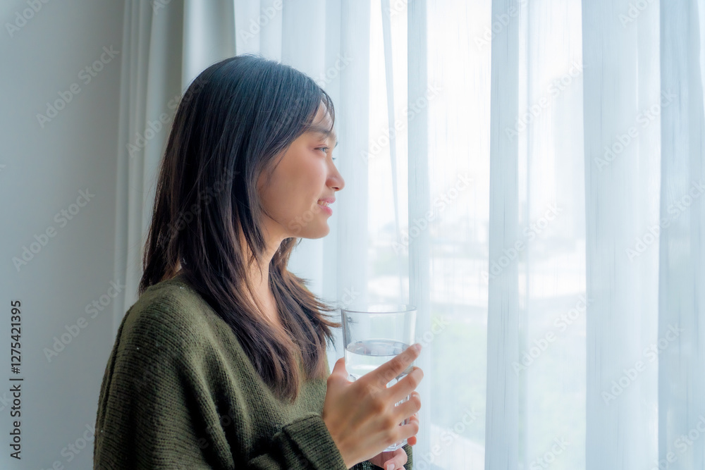 Close-up of Asian Woman with Brunette Ponytail Smiling While Drinking a Glass of Water in a White Room. Healthy Lifestyle, Hydration, and Wellness Concept. Close-up of Asian Woman with Brunette Ponytail Smiling While Drinking a Glass of Water in a White Room. Healthy Lifestyle, Hydration, and Wellness Concept.