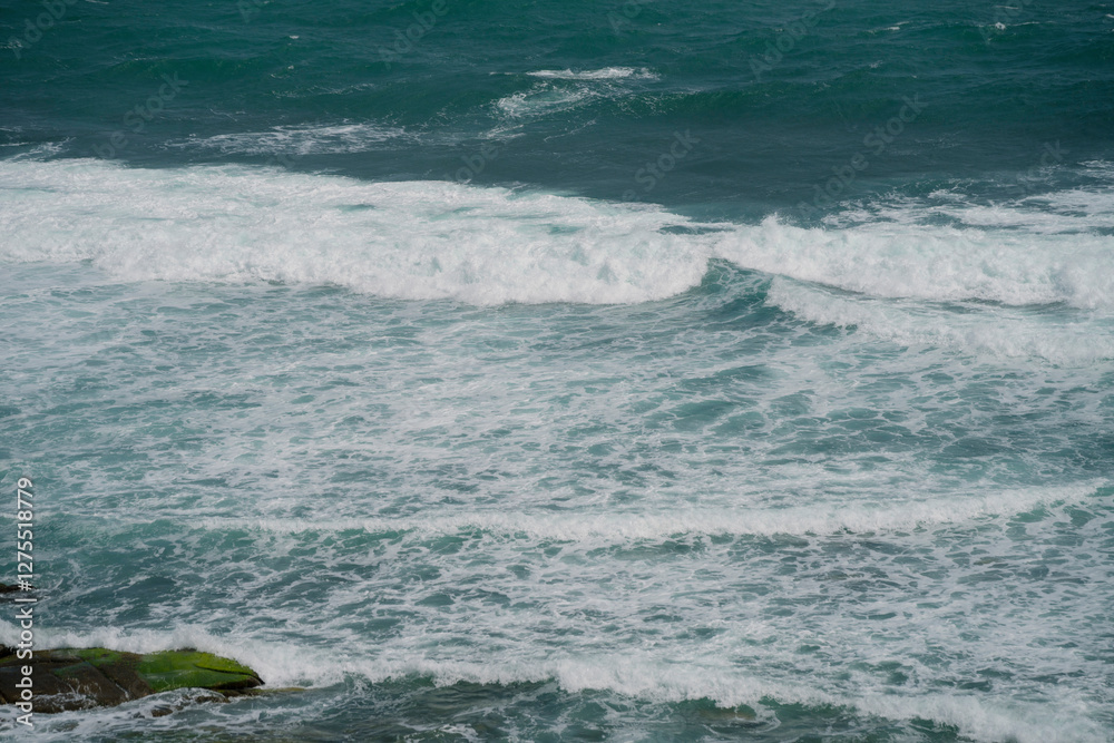 Fototapeta premium Powerful ocean waves crash against the shore, creating white foamy patterns on the deep blue sea. The swirling water and rhythmic waves reflect the beauty and energy of nature.