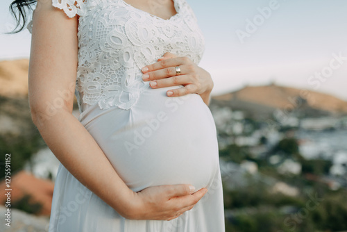 Pregnant woman in a white holiday dress hugs her belly against the background of mountains and the sea