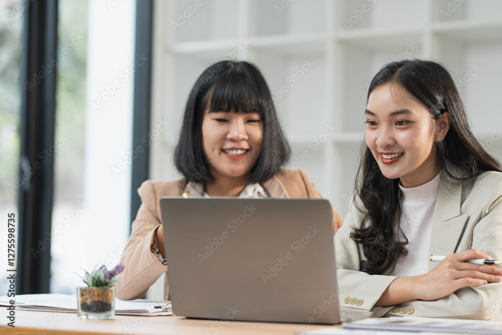 Two young businesswomen sitting at a modern office desk, collaborating on a project while using a laptop. Displaying teamwork and focus, they engage in productive discussions and strategic planning Two young businesswomen sitting at a modern office desk, collaborating on a project while using a laptop. Displaying teamwork and focus, they engage in productive discussions and strategic planning