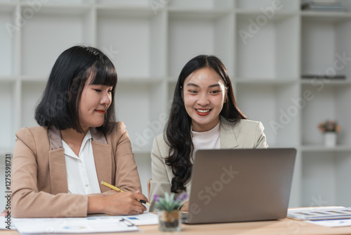 Two asian businesswomen are collaborating on a project, analyzing charts and reports on a laptop in a modern office, working together to achieve business success