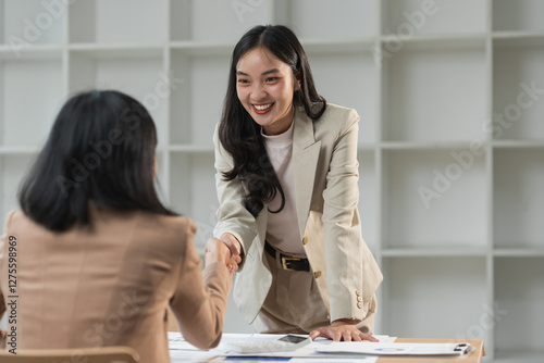 Two asian businesswomen shaking hands in the office, celebrating their successful collaboration after closing a deal and feeling proud of the results achieved together