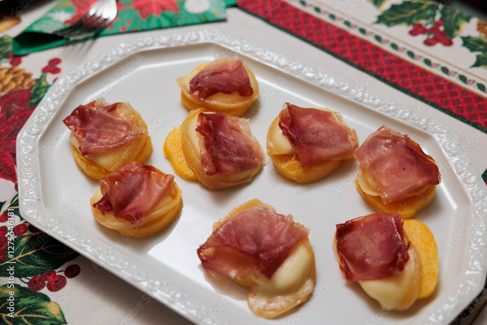dettaglio di un piatto visto dall'alto con degli antipasti a base di fette rotonde di polenta, scamorza affumicata e prosciutto crudo, su un tavolo in tema natalizio