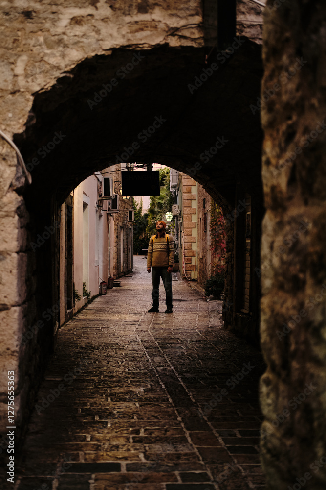 Obraz premium Narrow, atmospheric alley in an old European town Budva, framed by a rustic stone archway. A traveler with backpack stands in the soft morning light, gazing at the historic surroundings.