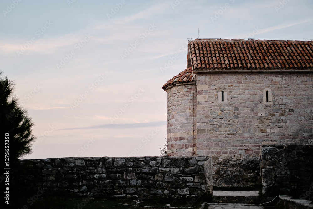 Fototapeta premium Ancient stone building with a red-tiled roof - small chapel or monastery. Pastel sky at sunrise adds a peaceful and timeless atmosphere. Old town Budva, Montenegro.