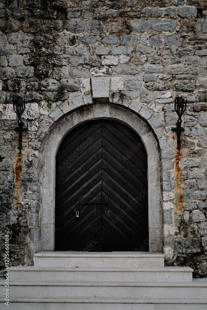 Black wooden door with a chevron pattern, set within an arched stone entrance. The textured stone wall and wrought-iron lanterns add a historic and elegant touch, while a padlock secures the door.