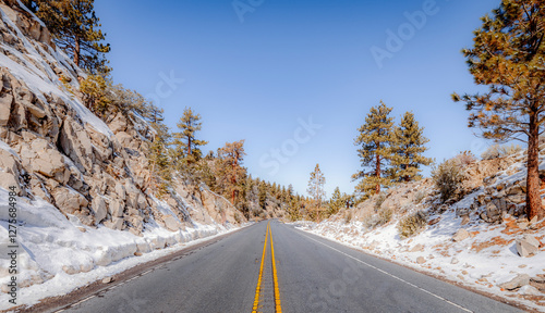 Icy road lined with snow cuts through a mountain valley with boulders and trees during mid day on the way to Big Bear Lake, CA 