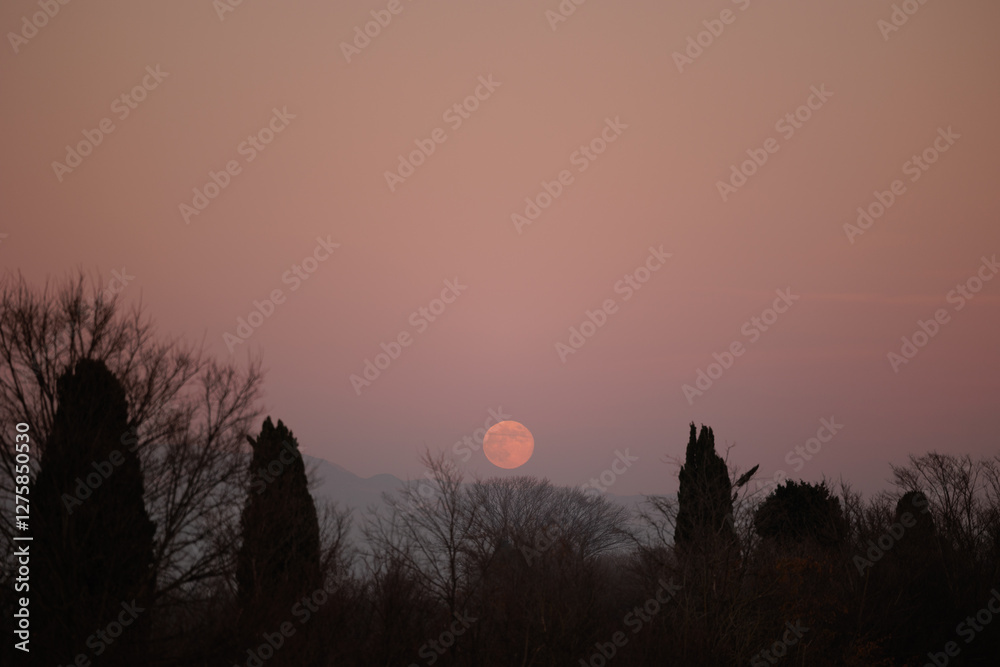 luna piena e di color arancione che sorge in un cielo sereno, dietro alle montagne tra Slovenia ed Italia, velata da una leggera foschia, in inverno, al tramonto