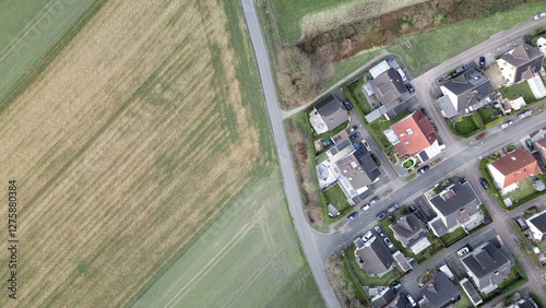 Aerial top-down view of a rural landscape with a paved road separating agricultural fields and a suburban residential area. A contrast between nature and urban development, showing farmland, houses, a