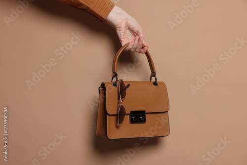 Woman showing stylish handbag and sunglasses on beige background, closeup photo