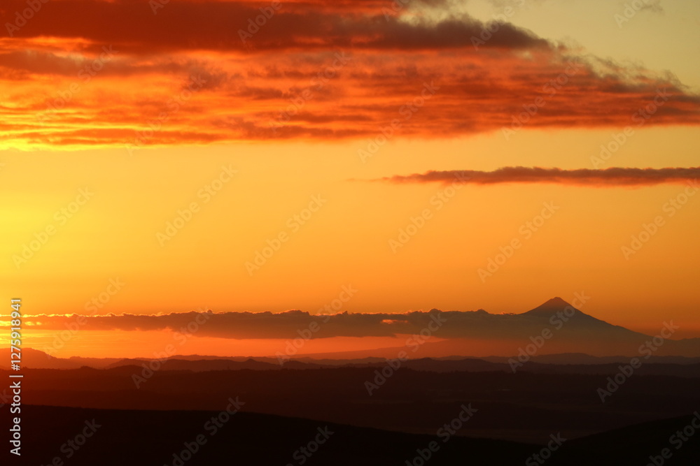 Obraz premium Fiery orange sky at sunset over a rugged mountain range in New Zealand. The vivid colors contrast with the darkening horizon, creating a mesmerizing natural spectacle.