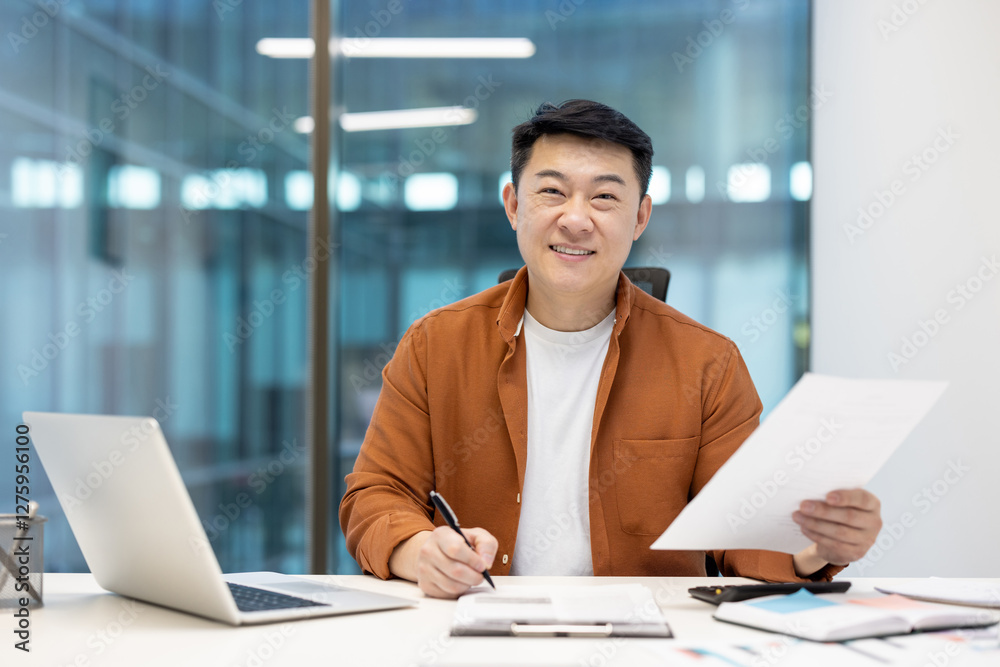 Portrait of successful Asian businessman financier at workplace inside office. Man behind paperwork looking at camera with satisfaction, working with laptop, preparing reports. Portrait of successful Asian businessman financier at workplace inside office. Man behind paperwork looking at camera with satisfaction, working with laptop, preparing reports.