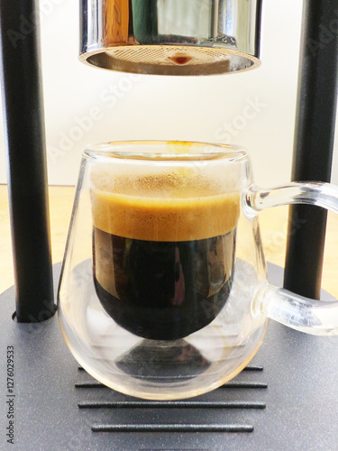 Closeup of a freshly poured hot espresso coffee with crema on top, in a glass cup, standing on the base of the manual machine which extracted the shot. photo