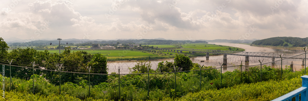 Panoramic View Of Lush Green Fields And Winding River With Bridges And Cable Cars Beyond A Barbed Wire Fence Panoramic View Of Lush Green Fields And Winding River With Bridges And Cable Cars Beyond A Barbed Wire Fence