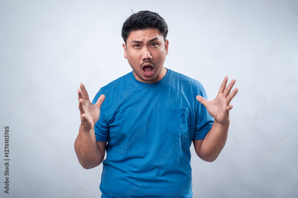 Fototapeta premium An Asian man in a blue t-shirt stands against a plain white background, raising both hands in a surprised and frustrated expression. His facial reaction conveys shock, disbelief, or confusion.