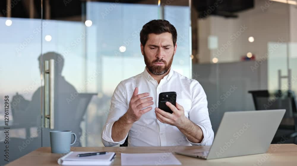 Frustrated businessman trying to get better mobile network at office. Bearded male sitting at desk looking at smartphone raising arm with bad connection.