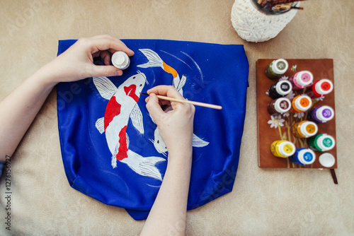 Female artist sitting on the couch and painting on fabric koi carp. Customization of clothes with acrylic paints on fabric. Custom made and bespoke clothes