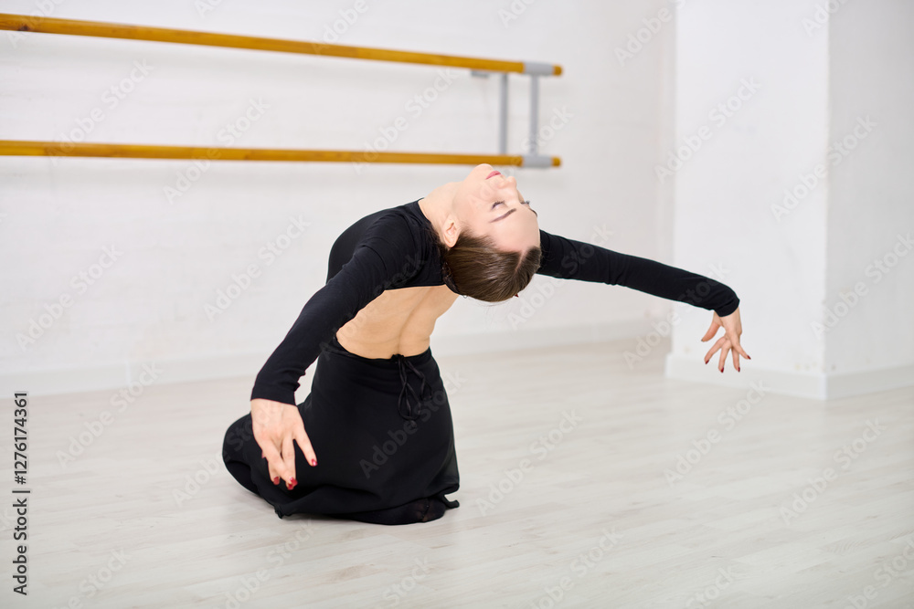 Naklejka premium Young female ballerina with brown hair kneels on studio floor, arching back with arms extended. Soft lighting illuminates black leotard light wooden floor visible