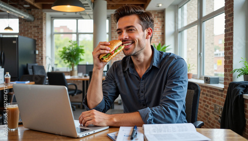 Smiling man enjoying a sandwich at a modern office desk