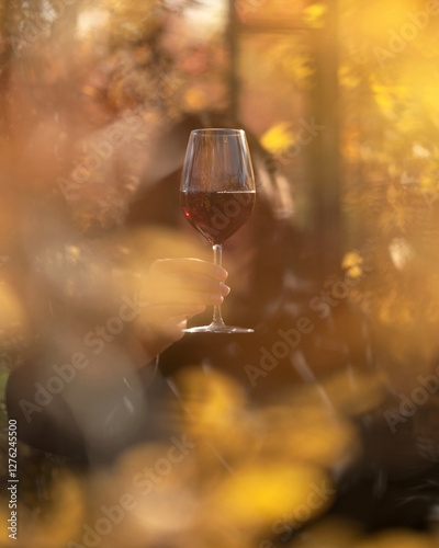 Glass of red wine in hand in autumn. Wineglass. Wine tasting. Aperitif. Alcohol beverage. Ruby wine. Drink concept. Blurred golden foliage, fall season. Copy space. Warm atmosphere, golden bokeh.