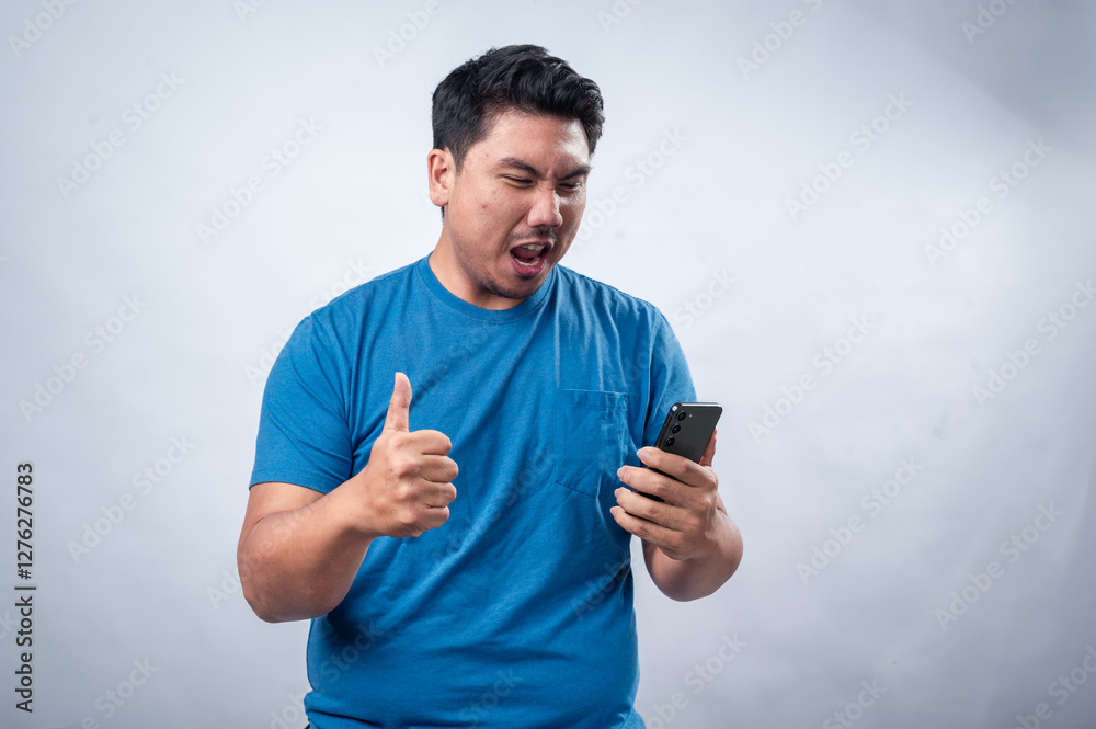 An Asian man wearing a blue t-shirt holds a smartphone in one hand while giving a thumbs-up with the other. He smiles confidently against a plain white isolated background An Asian man wearing a blue t-shirt holds a smartphone in one hand while giving a thumbs-up with the other. He smiles confidently against a plain white isolated background