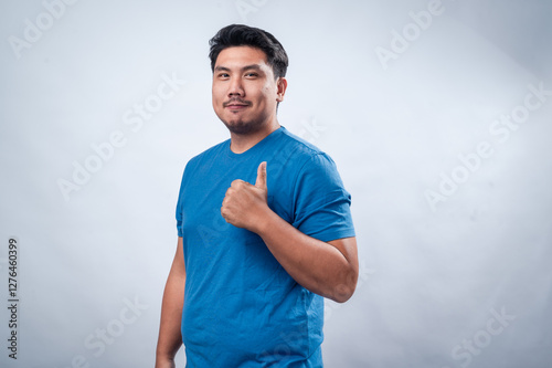 A cheerful Asian man wearing a blue t-shirt gives a thumbs-up gesture while smiling, standing against a clean white background. His positive expression and body language convey confidence and approval photo