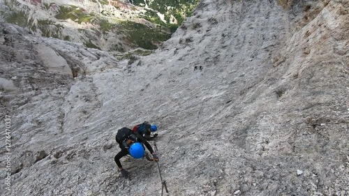Two intrepid climbers ascend a steep, rocky mountain face, their figures small against the vast, textured landscape under a soft, diffused sunlight.