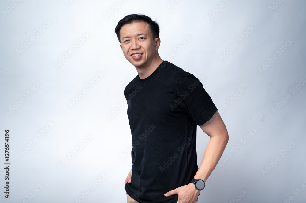 An Asian man in a black t-shirt and beige pants is smiling confidently while posing with his hands in his pockets. He is wearing a black smartwatch on his wrist. The background is plain white An Asian man in a black t-shirt and beige pants is smiling confidently while posing with his hands in his pockets. He is wearing a black smartwatch on his wrist. The background is plain white