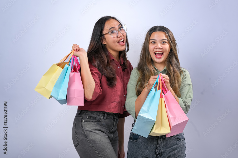 Two young Asian women with long hair wear casual crop shirts, one red and one green, with dark jeans. They hold colorful shopping bags and smile excitedly while posing together against a plain white Two young Asian women with long hair wear casual crop shirts, one red and one green, with dark jeans. They hold colorful shopping bags and smile excitedly while posing together against a plain white