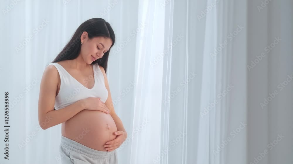 Young happy Asian pregnant woman posing in studio with white curtains in background Young happy Asian pregnant woman posing in studio with white curtains in background