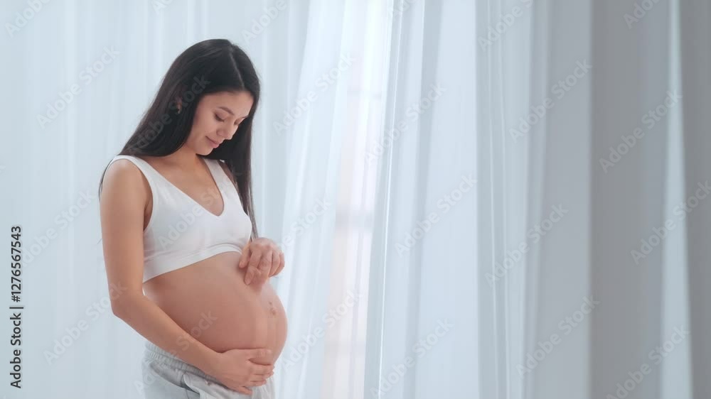 Young happy Asian pregnant woman posing in studio with white curtains in background Young happy Asian pregnant woman posing in studio with white curtains in background