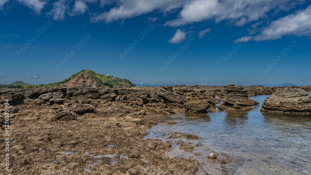 Fototapeta premium Low tide in the ocean. Exposed boulders of the seabed are scattered in shallow water. A tiny silhouette of a man on a rocky coast, Blue sky, clouds.Madagascar. Nosy Be