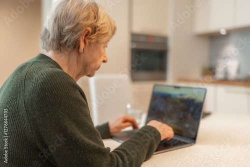 Senior woman using laptop at home, looking at the screen and typing on keyboard. Concept of technology, online shopping, digital literacy for seniors and active aging. Technology usage among seniors. photo