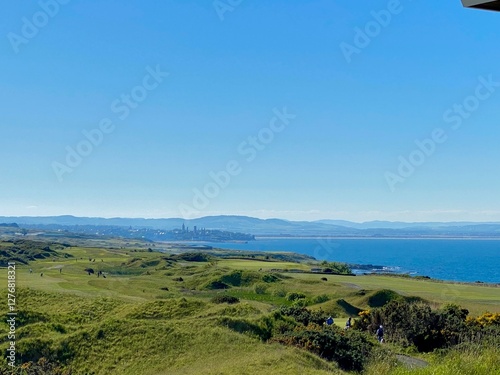 golf course with coastal view. Distant city skyline and blue sea. Green grass and rolling hills. Sunny day with clear sky. Outdoor sport and travel.