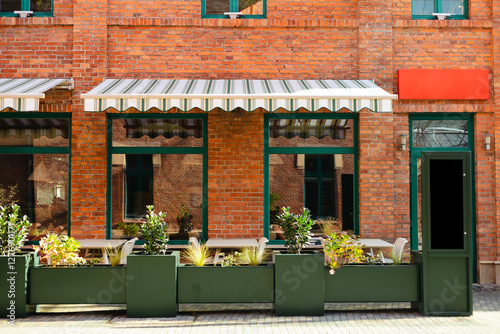 Street cafe with brick walls and canopy exterior design. Front view, sunny day. An outdoor cafe with tables, chairs. Lush green plants growing in pots near house. High quality photo.