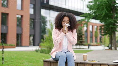 A sick African American female student suffering from runny nose and cold itting on a bench near the university building. Young black woman wipes her nose with a handkerchief. She has virus or flu photo