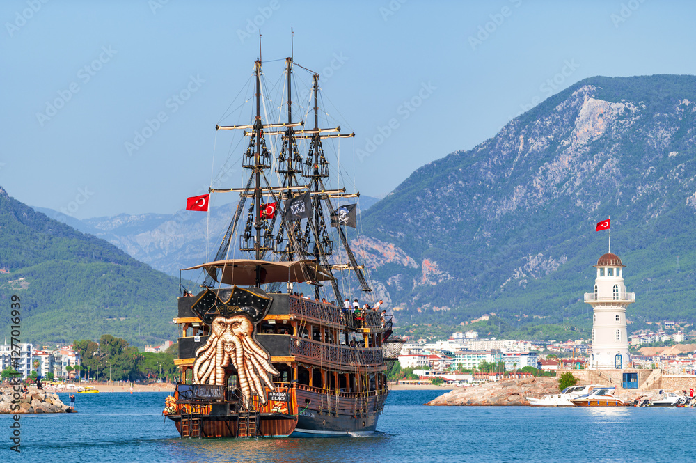 Alanya, Antalya, Turkey. 15 july 2023. Pirate ship sails into harbor, a captivating blend of history and tourism with a lighthouse backdrop and mountains far beyond. Coastal scenery Alanya, Antalya, Turkey. 15 july 2023. Pirate ship sails into harbor, a captivating blend of history and tourism with a lighthouse backdrop and mountains far beyond. Coastal scenery