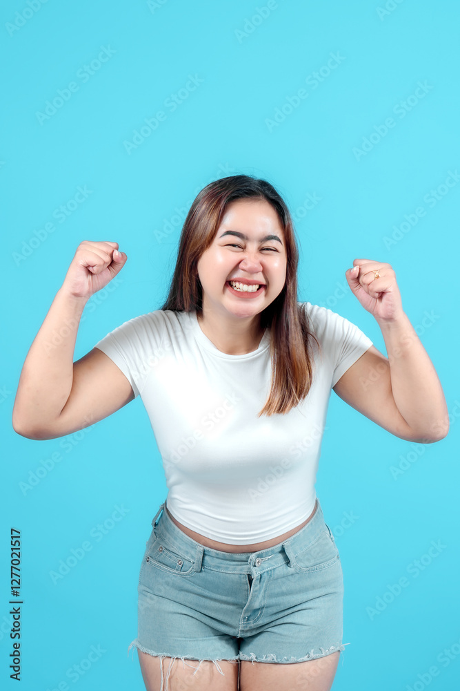 A joyful young woman celebrating with raised fists against a blue background, expressing happiness and excitement. Perfect for themes of success, positivity, and achievement. A joyful young woman celebrating with raised fists against a blue background, expressing happiness and excitement. Perfect for themes of success, positivity, and achievement.