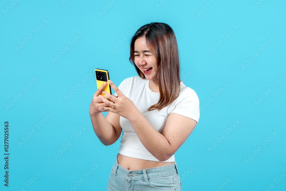 A young woman in a white t-shirt joyfully looks at her yellow smartphone, smiling widely against a bright blue background, capturing a moment of happiness and engagement A young woman in a white t-shirt joyfully looks at her yellow smartphone, smiling widely against a bright blue background, capturing a moment of happiness and engagement