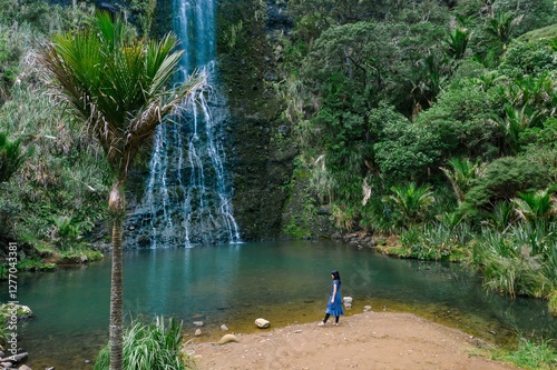 Woman in a blue dress walks along the edge of a tranquil waterfall pool, surrounded by lush greenery. Peaceful nature scene. Karekare Falls, Karekare, Auckland, New Zealand photo